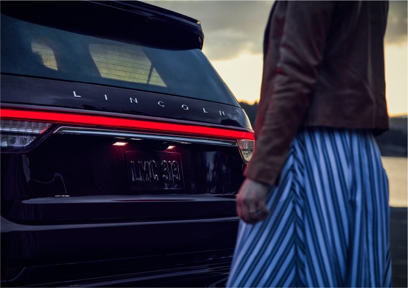 A person is shown near the rear of a 2023 Lincoln Aviator® SUV as the Lincoln Embrace illuminates the rear lights | Lincoln of Coconut Creek in Coconut Creek FL