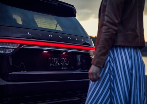 A person is shown near the rear of a 2024 Lincoln Aviator® SUV as the Lincoln Embrace illuminates the rear lights | Lincoln of Coconut Creek in Coconut Creek FL